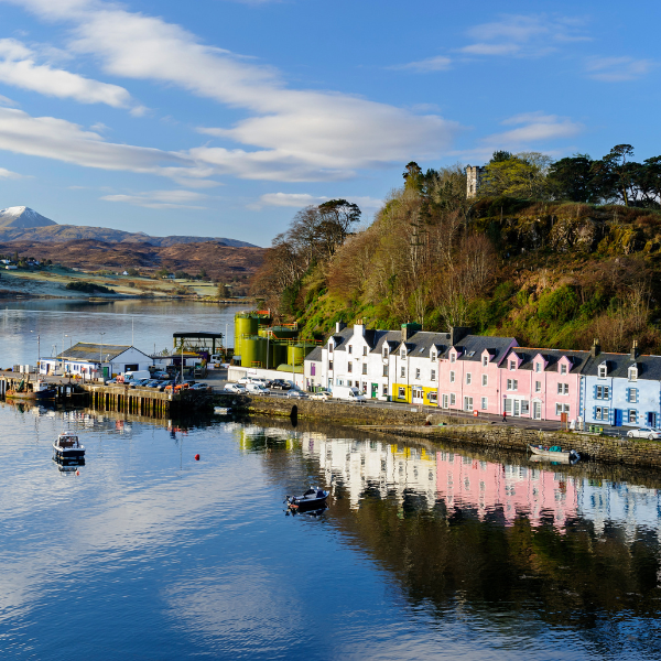 A row of colourful terraced houses by the sea, next to a small harbour. There are snow capped mountains in the background and a blue cloudy sky.